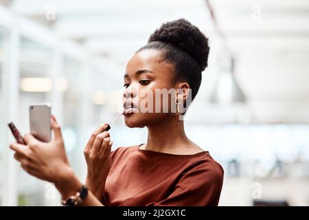 Make-up macht mich zum Chef fühlen. Aufnahme einer jungen Geschäftsfrau, die Lippenstift anwendet, während sie ihr Mobiltelefon als Spiegel in einem Büro benutzt. Stockfoto