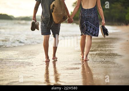 Spaziergang bei Sonnenuntergang. Zugeschnittenes Bild eines nicht identifizierbaren Paares, das sich beim Strandspaziergängen die Hände hält. Stockfoto