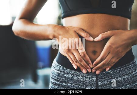 Der Alltag ist der Tag der Kernstärkung. Eine kurze Aufnahme einer fittenden jungen Frau, die in einem Fitnessstudio eine herzförmige Geste über ihren Bauch macht. Stockfoto