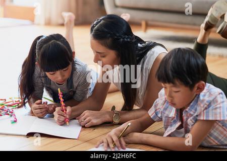 Das ist, wie es gemacht hat, meine Liebe. Aufnahme einer fröhlichen Mutter und ihrer beiden Kinder, die tagsüber gemeinsam Hausaufgaben machen, während sie zu Hause auf dem Boden liegen. Stockfoto