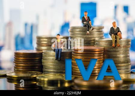 Miniaturmenschen: Kleine Geschäftsleute, die auf Münzstapeln sitzen. Steuerkonzept. Die Buchstaben IVA (Spanish Value Added Tax) auf Würfel auf Haufen von Gold CO Stockfoto