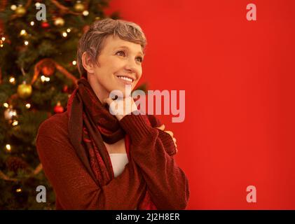 Es ist die Jahreszeit, in der es richtig fröhlich ist. Eine glückliche reife Frau, die nachdenklich vor einem Weihnachtsbaum aussieht. Stockfoto
