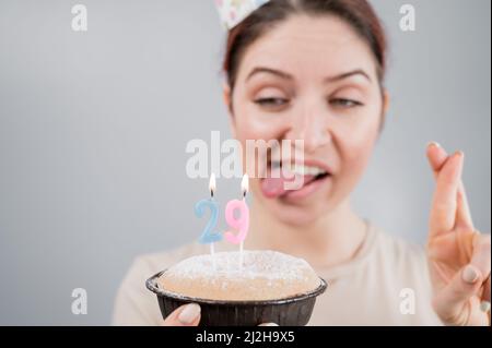 Glückliche kaukasische Frau, die ihre Zunge herausstreckte und mit gekreuzten Fingern die Kerzen auf dem Kuchen ausbläst. Das Mädchen feiert ihren 29. Geburtstag Stockfoto