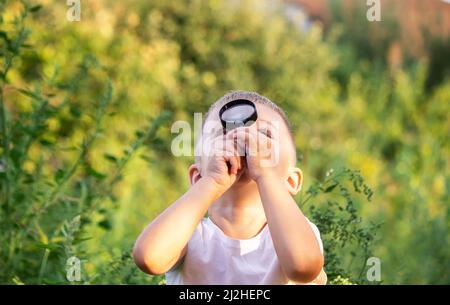 Kleiner Junge, der die Natur auf der Wiese mit einer Lupe erkundet und Blumen betrachtet. Neugierige Kinder im Wald, ein zukünftiger Botaniker. Stockfoto