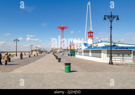 Roter Fallschirmsprung des Vergnügungsparks Luna Park mit Strandpromenade vor dem Hotel, Spaziergänger, Straßenlaternen und grünen Mülltonnen in einer Reihe, Coney isran Stockfoto