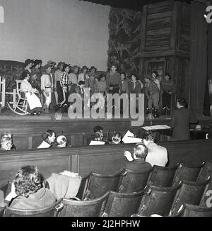 1960s, historische, große Gruppe von Schauspielern auf einer Bühne während einer Aufführung des amerikanischen Musicals Oklahoma, produziert von der Kelty Musical Association in der Carnegie Hall, Dufermline, Fife, Schottland, Großbritannien, Mit Orchester in der Grube. Oklahoma basiert auf dem Theaterstück „Green Grow The Lilacs“ aus dem Jahr 1931 und ist das erste Musical von Rodgers und Hammerstein, das 1943 am Broadway eröffnet wurde. Basierend auf den Romanzen eines einfachen Bauernmädchen ist das Musical eine beliebte Wahl für Schulen und Gemeinschaftsgruppen. Stockfoto