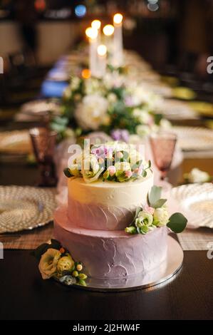 Weißer Hochzeitstorte, geschmückt mit Blumen auf einem servierten Tisch. Schwarzer Hintergrund eines Restaurants. Abendgesellschaft, Feier. Stockfoto