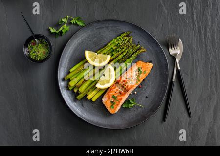 Gebratenes Lachsfilet mit grünem Spargel, serviert mit Zitronenvinaigrette auf dunklem Schiefergrund Stockfoto