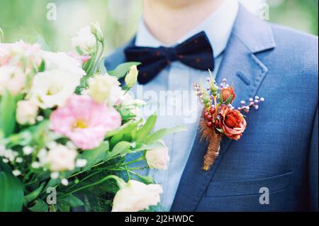 Hochzeitsdetails aus nächster Nähe. Bräutigam in blauem Anzug, trägt Boutonniere mit roten Blumen und hält einen Brautstrauß Stockfoto