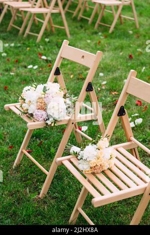 Hochzeitszeremonie im Freien. Blumensträuße der Brautjungfer mit zarten Blumen auf Holzstühlen auf grünem Gras. Stockfoto
