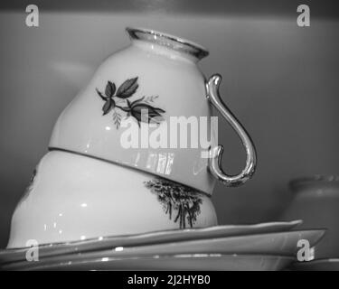 Teetassen in Schwarz und Weiß, Vintage altmodische antike Teetassen und Untertassen, hübsches Blumendesign, gestapelt auf einem Küchenschrankregal, monochrom Stockfoto