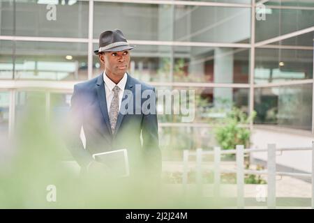 Porträt eines Gentleman mit einem Bürogebäude im Hintergrund. Junger Geschäftsmann in Anzug, Krawatte und Hut, der ein digitales Tablet in der Hand hält. Stockfoto