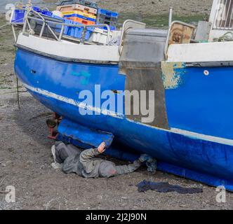 Mann, der bei Ebbe auf dem Rücken liegt und den Fiberglas-Bootsrumpf reinigt Stockfoto