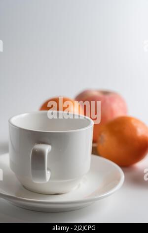 Nahaufnahme einer weißen Tasse und Untertasse mit Orangen und einem Apfel im Hintergrund Stockfoto