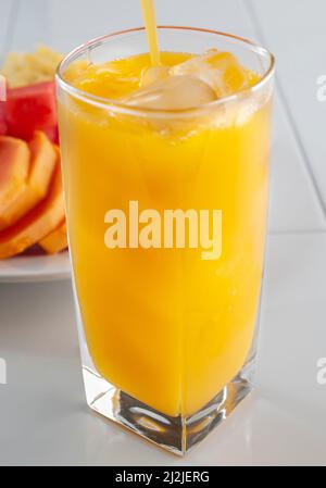 Ein Mango-Saft in einem quadratischen Glas mit Eis, auf einer weißen Küchenoberfläche und einem Teller mit geschnittenen Früchten auf dem Hintergrund. Vertikales Bild Stockfoto