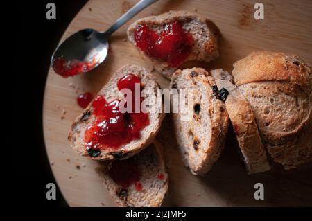 Brotscheiben mit Rosinen und Erdbeermarmelade oben, mit einem Löffel an der Seite, auf einem hölzernen Schneidebrett aus der Nähe von oben, Food-Fotografie Stockfoto