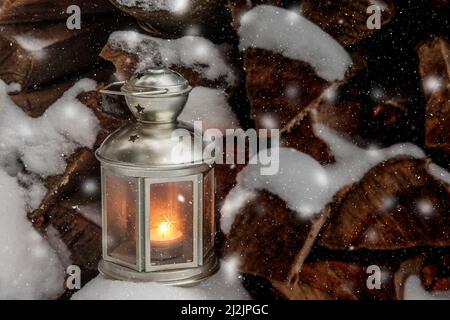 Die Weihnachtslaterne mit brennender Kerze steht auf einem verschneiten Holzstapel und es beginnt sanft zu schneien. Stockfoto