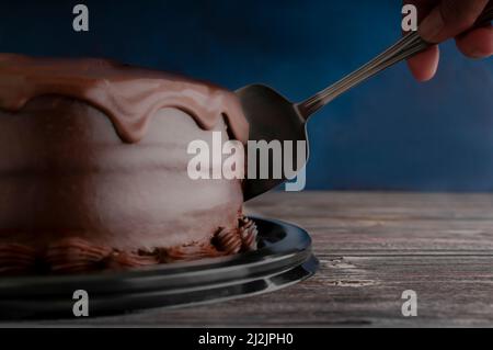 Eine Hand, die einen Schokoladenkuchen auf einem Holztisch mit dunkelblauem Hintergrund schneidet, Fotos von dunklen Speisen Stockfoto