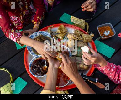 Draufsicht auf die Hände der muslimischen Familie, die am Ende des Ramadan Mubarak auf einem runden Tablett auf einem schwarzen Tisch eine Vielzahl traditioneller malaiischer Gerichte genießen Stockfoto