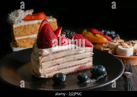 Scheibe Bananenkrepskuchen mit Creme und Erdbeeren oben mit blauen Beeren auf einem schwarzen Teller. Makro, dunkle Food-Fotografie. Stockfoto