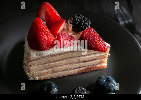 Scheibe Bananenkrepskuchen mit Creme und Erdbeeren oben mit blauen Beeren auf einem schwarzen Teller. Makro, dunkle Food-Fotografie. Stockfoto