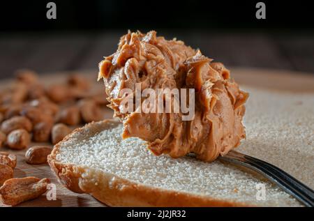 Makrofotografie. Erdnussbutter in einem Löffel auf einer Brotscheibe, mit Erdnüssen im Hintergrund, auf einem Holzschneidebrett, Stockfoto