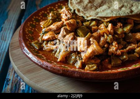 Schweinefleisch. Mexikanisches Essen, Huhn mit Nopales und handgemachte Maistortillas auf einem Schlamm- oder Tonteller, auf einem runden Holzschneidebrett, Stockfoto