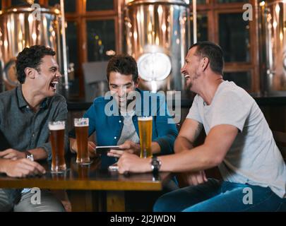 Es ist Zeit, bei einem Bier über das Geschäft zu sprechen. Aufnahme von drei fröhlichen jungen Männern, die tagsüber gemeinsam an einem Tisch in einer Bierbrauerei Bier trinken. Stockfoto