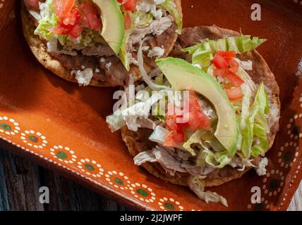 Traditionelle mexikanische Speisen Tostadas auf einer Lehm- oder Schlammplatte über einem rustikalen Tisch mit Huhn oder Carnitas, Salat, Tomaten, Zwiebeln, gebratenen Bohnen, Würzig Stockfoto