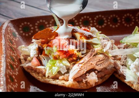 Traditionelle mexikanische Speisen Tostadas auf einer Lehm- oder Schlammplatte über einem rustikalen Tisch mit Huhn oder Carnitas, Salat, Tomaten, Zwiebeln, gebratenen Bohnen, Würzig Stockfoto