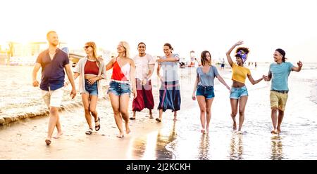 Große Gruppe von trendigen, fröhlichen Freunden, die bis zum Sonnenuntergang am Strand spazieren gehen - multirassisches Lifestyle-Konzept mit jungen Leuten, die Spaß im Sommerurlaub haben - Stockfoto