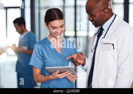 Sie haben heute um drei Uhr einen Termin. Aufnahme von zwei fröhlichen jungen Ärzten, die über ein digitales Tablet im Inneren eines Krankenhauses diskutieren. Stockfoto