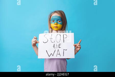 Ein trauriges Mädchen mit einem gespaltenen Gesicht in den Farben der ukrainischen Flagge hält ein Antikriegsplakat auf einem blau isolierten Hintergrund. Es gibt keinen Krieg im Koncep Stockfoto