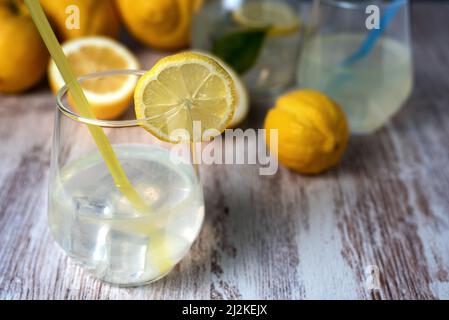 Glas mit Limonade und Eis, mit einer Scheibe Zitrone am Rand, im Hintergrund ein Krug und einige Zitronen aus dem Fokus. Stockfoto