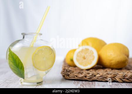 Herzförmiger Krug mit Limonade und Würfeln, einer Zitronenscheibe und einem grünen Blatt, im Hintergrund einige Zitronen unscharf. Stockfoto