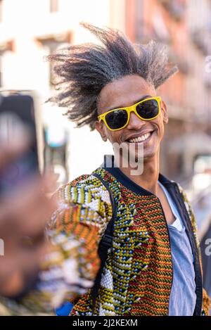 Hübscher junger Mann, der sein Smartphone für ein Selfie, lässige Kleidung und Street-Wear, coole Attitüde und afro amerikanische Mischvolk benutzt Stockfoto
