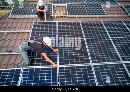 Männer Installateure installieren Photovoltaik-Solarmodule auf dem Dach des Hauses. Ingenieure in Helmen bauen Solarpanel-System im Freien. Konzept der alternativen und erneuerbaren Energien. Luftaufnahme. Stockfoto