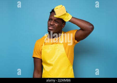 Müde junger afrikanischer Mann trägt gelbe Gummihandschuhe zum Schutz der Hände und wischt Schweiß ab, während er das Haus auf blauem Hintergrund isoliert putzt Stockfoto