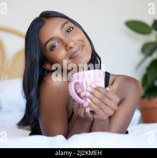 Eine frische Tasse Kaffee am Morgen. In voller Länge Porträt einer attraktiven jungen Frau, die eine Tasse Kaffee genießt, während sie auf einem Bett liegt. Stockfoto