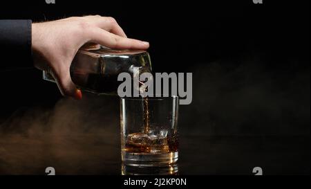 Der Barkeeper gießt Whiskey Cognac-Brandy aus der Flasche in das Trinkglas mit Eiswürfeln auf dunklem Hintergrund Stockfoto