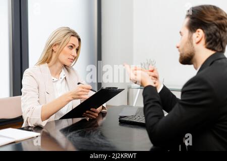 Helle Idee. Fröhliche junge qualifizierte Kollegen sitzen im Büro am Tisch und diskutieren mit einem Lächeln über ihr Projekt. Die Frau hält Dokumente, während der Mann positiv gestikiert Stockfoto