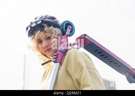 Schöne blonde kaukasische Frau in einem Helm und gelben Pullover, hält einen rosa Roller auf ihrer Schulter außerhalb weißen Hintergrund Porträt . Hochwertige Fotos Stockfoto