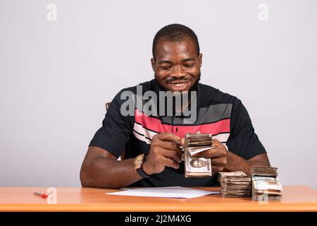 Ein junger glücklicher schwarzer Geschäftsmann in einem T-Shirt, der an einem Tisch sitzt und Banknoten zählt Stockfoto