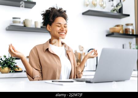 Hübsche, positive, ruhige, entspannte afroamerikanische junge Frau, Studentin, Freiberuflerin oder Designerin, die fernab von zu Hause an einem Laptop arbeitet, sich ausruhen und meditieren während der Arbeit, geschlossene Augen, ruhiges Konzept Stockfoto