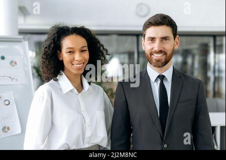 Portrait von zwei erfolgreichen Kollegen aus verschiedenen Rassen, Firmengründern, Direktoren, Managern, einem kaukasischen Mann und einer afroamerikanischen Frau, die in einem modernen Büro stehen und lächelnd auf die Kamera blicken Stockfoto