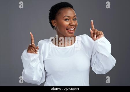 Ich bin so aufgeregt und ich kann es einfach nicht verbergen. Aufnahme einer jungen Frau, die vor einem grauen Hintergrund nach oben zeigt. Stockfoto