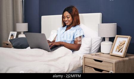 Sie arbeitet immer dann auf ihrem Blog, wenn sie die Chance hat. Eine kurze Aufnahme einer attraktiven jungen Frau, die ihren Laptop benutzt, während sie im Bett sitzt. Stockfoto