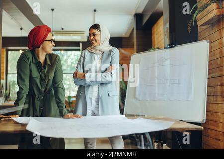 Fröhliche weibliche Designerinnen, die während der Arbeit an Blaupausen-Zeichnungen diskutieren. Zwei kreative Geschäftsfrauen planen ein neues Projekt in einem Büro. Lichtbogen Stockfoto
