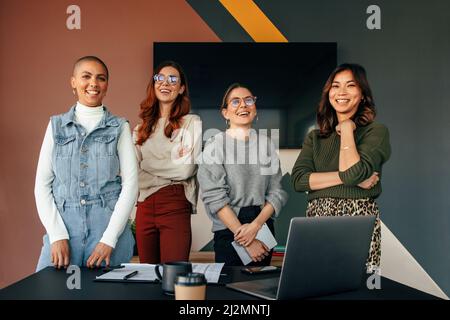 Erfolgreiche Unternehmerinnen lächeln fröhlich, während sie hinter einem Tisch in einem Sitzungssaal stehen. Gruppe von multikulturellen Geschäftsfrauen, die zusammenarbeiten Stockfoto