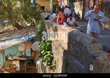 Eine Szene am heiligen Wallfahrtsort Banganga Tank in Walkeshwar, Mumbai, Indien, ein Mann, der im Schatten einer Hütte schläft, während ein Mann in der Nähe einen Snack genießt Stockfoto